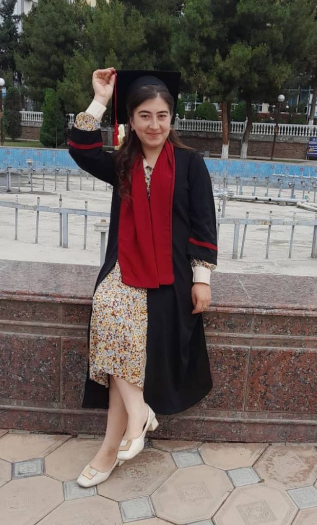 Young Central Asian woman in a dark graduation gown and cap with a red sash and tassel outside near leafy trees. 