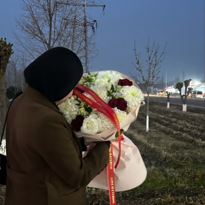 Person in a long brown coat and hood holding a large bouquet of red and white roses standing in a field of trees that are just budding.