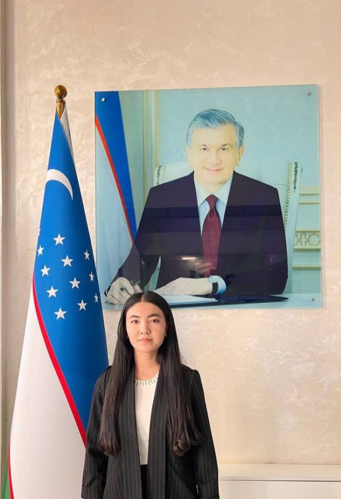 Young Central Asian woman with long dark hair and a long dark coat over a white top. Painting of a man in a suit and a flag near her.