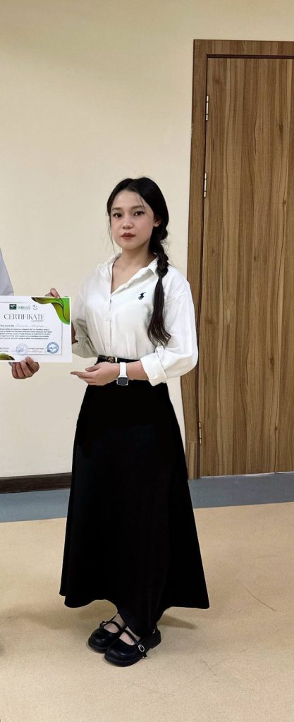 Young Central Asian woman with long straight dark hair in ponytail, a white collared shirt and wristwatch and black skirt, holding a certificate. 