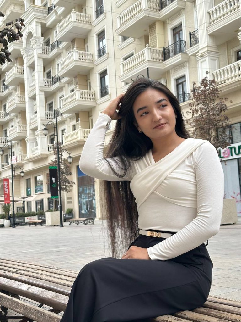 Young Central Asian woman with long straight dark hair and a white top in front of a large white apartment building with balconies. 