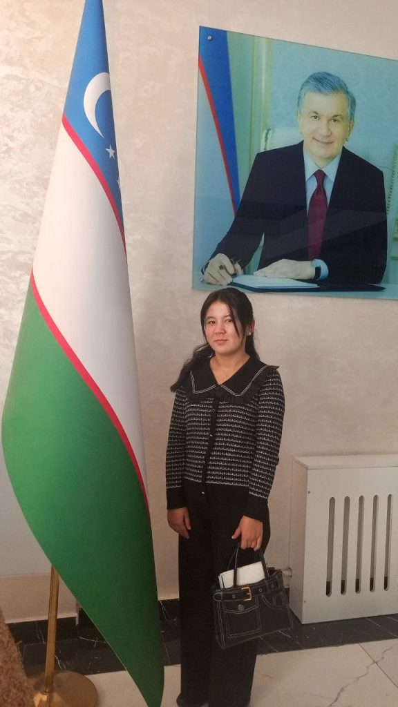 Young Central Asian woman with a black and gray striped sweater and black pants and long dark hair posing in front of a painting of a distinguished gentleman in a suit and standing near a flag. 