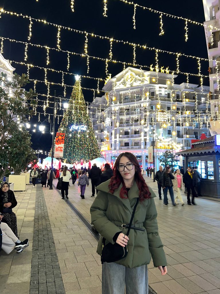 Young Central Asian woman with long dark curly hair, reading glasses, a green poofy jacket, and a black purse. She's outdoors at night near lit-up Christmas trees and lights suspended from poles. 