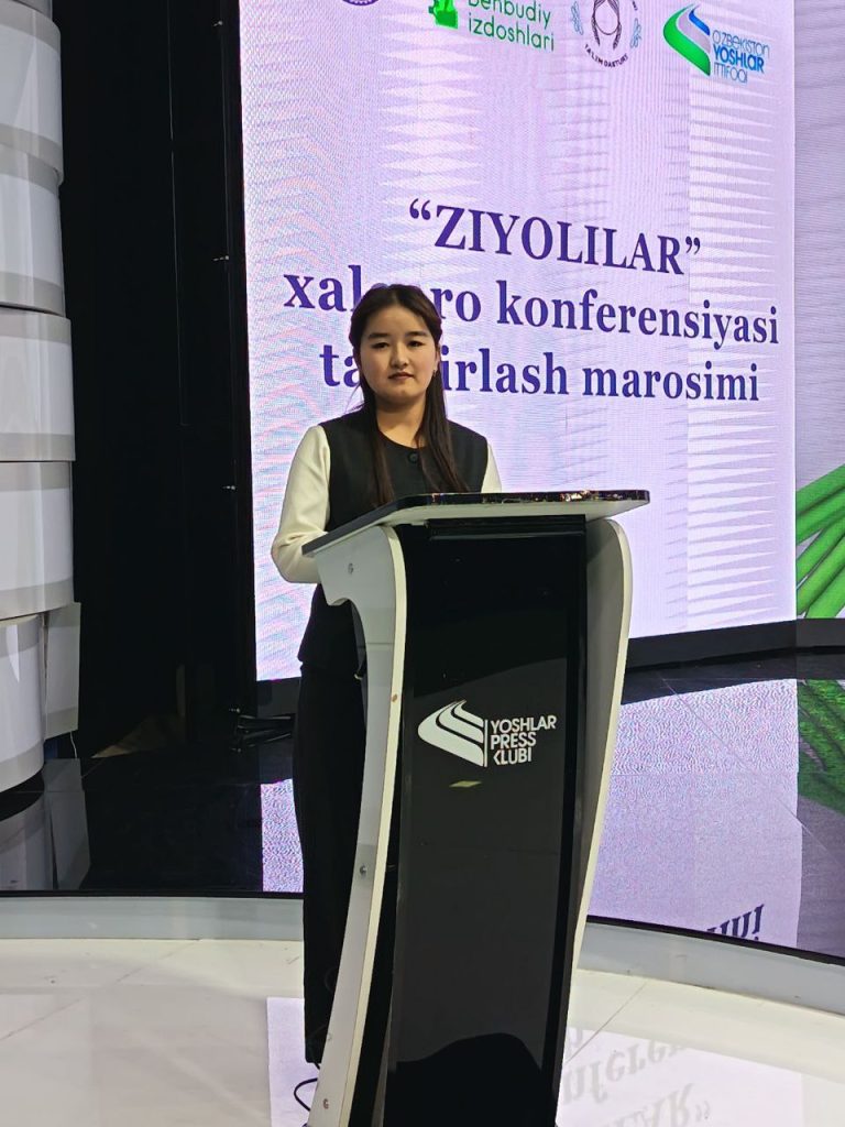 Young Central Asian woman with long dark straight hair, a black vest over a white top, standing at a lectern. 
