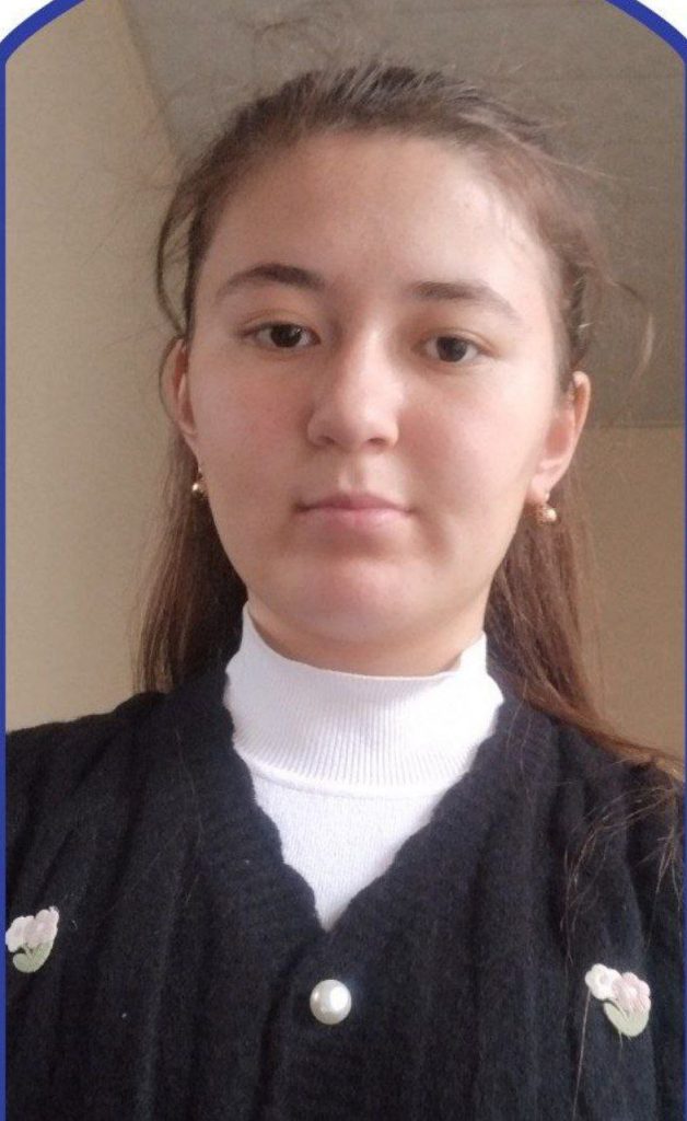 (Young Central Asian woman with long dark hair, brown eyes, earrings, and a black sweater over a white top)