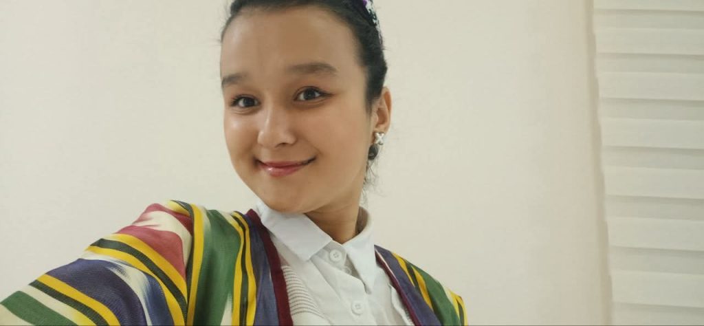 Young Central Asian woman with long dark hair up in a bun and a yellow and red and blue and green jacket over a white collared top. 