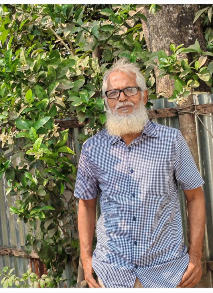 Older South Asian man with a mustache and beard, white hair, and reading glasses and a blue and white collared top standing outside near a leafy tree. 