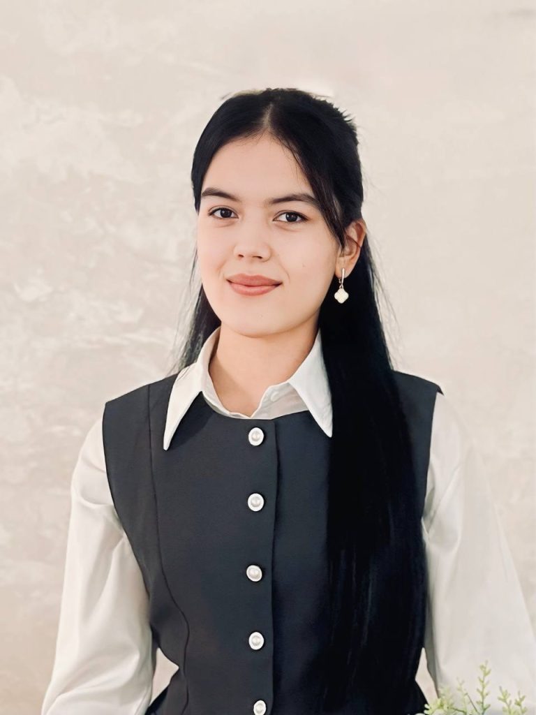Young Central Asian woman with long dark hair, brown eyes, small pendant earrings, and a black vest over a white collared shirt. 