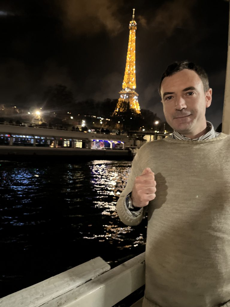 Young middle aged French man with short dark hair and a tan sweater standing out at night near the Seine River and the Eiffel Tower. 