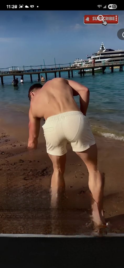 Young white man with no shirt and white shorts bends down at the beach near a cruise ship and pier. His back is to us. 