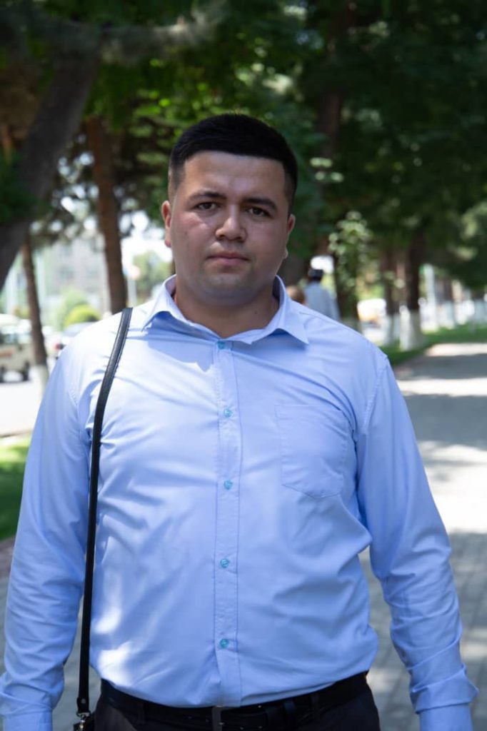 Young Central Asian man with short dark hair and a white collared shirt outside under leafy trees on a sunny day. 