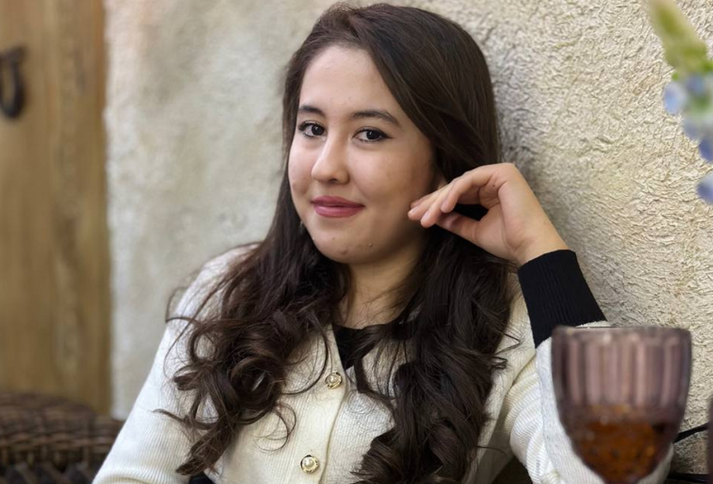 Young Central Asian woman with long dark curly hair, brown eyes, a white coat and black top, posing near a water glass.